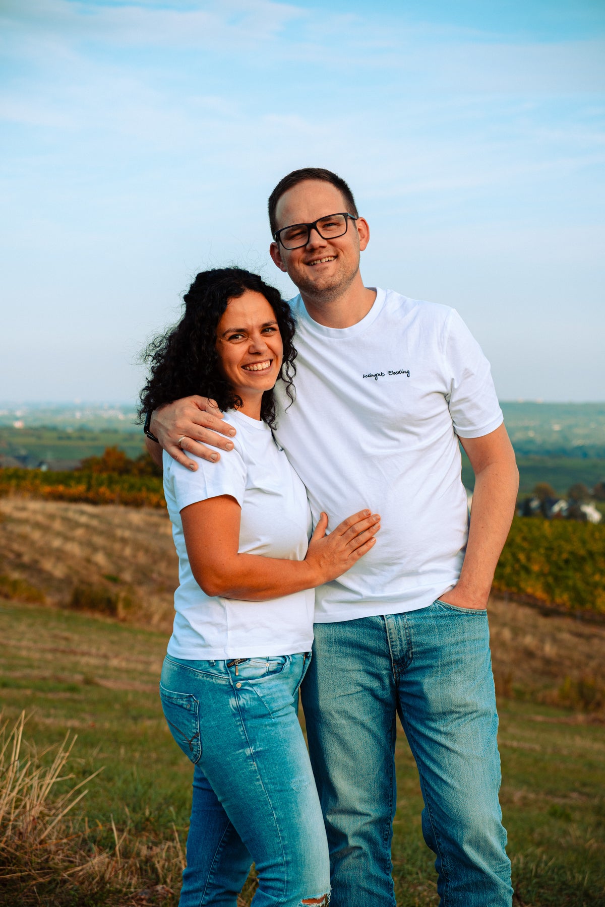 Paar Arm in Arm in weißen T-Shirts, auf seinem der gestickte Schriftzug „Weingut Eberling“, auf einer Wiese mit Weinbergen und weitem Blick ins Land