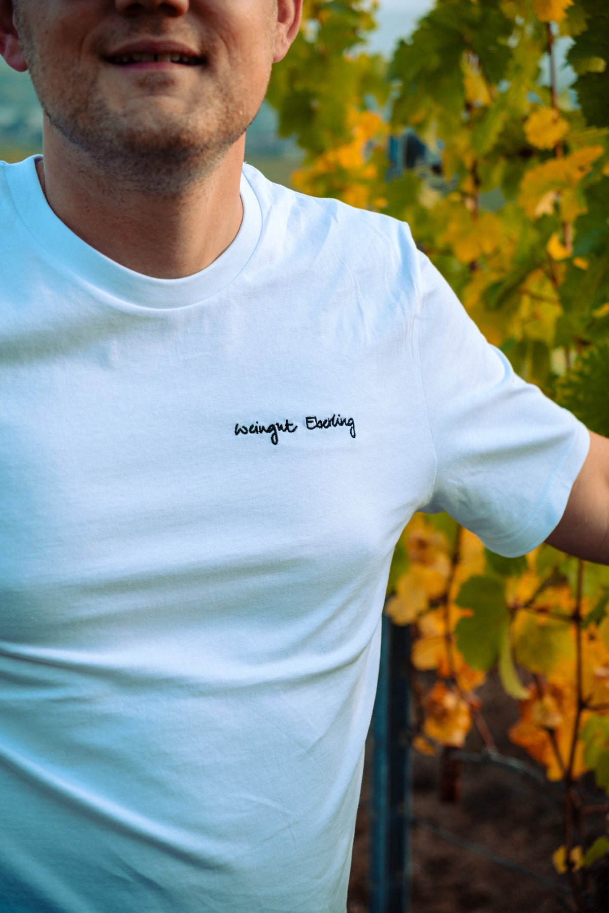 Weißes T-Shirt mit schwarz gesticktem Schriftzug „Weingut Eberling“ auf der linken Brust, getragen vor herbstlichem Weinlaub