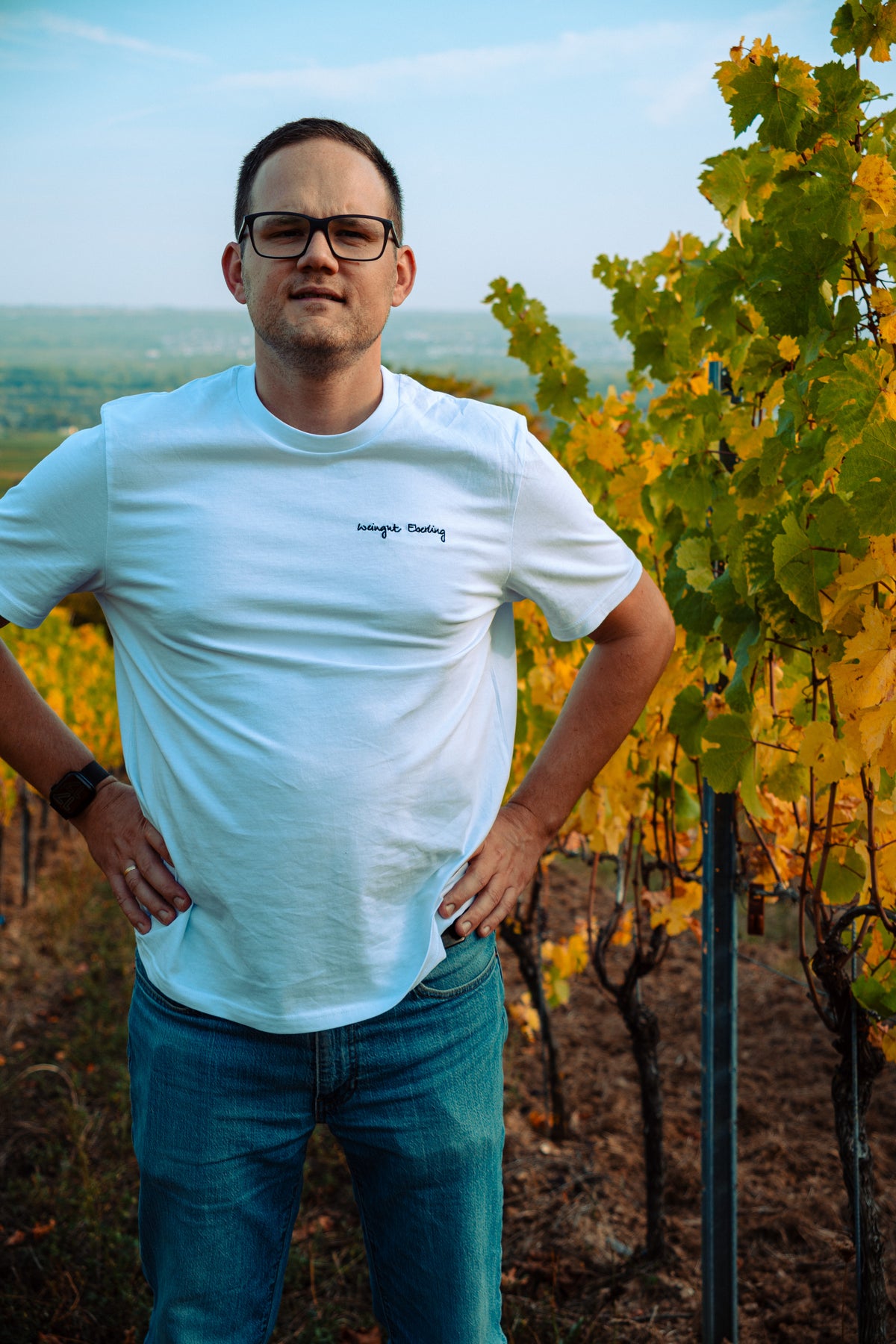 Mann mit Brille im weißen T-Shirt mit schwarz gesticktem Schriftzug „Weingut Eberling“ auf der linken Brust, Hände in den Hüften, im herbstlichen Weinberg