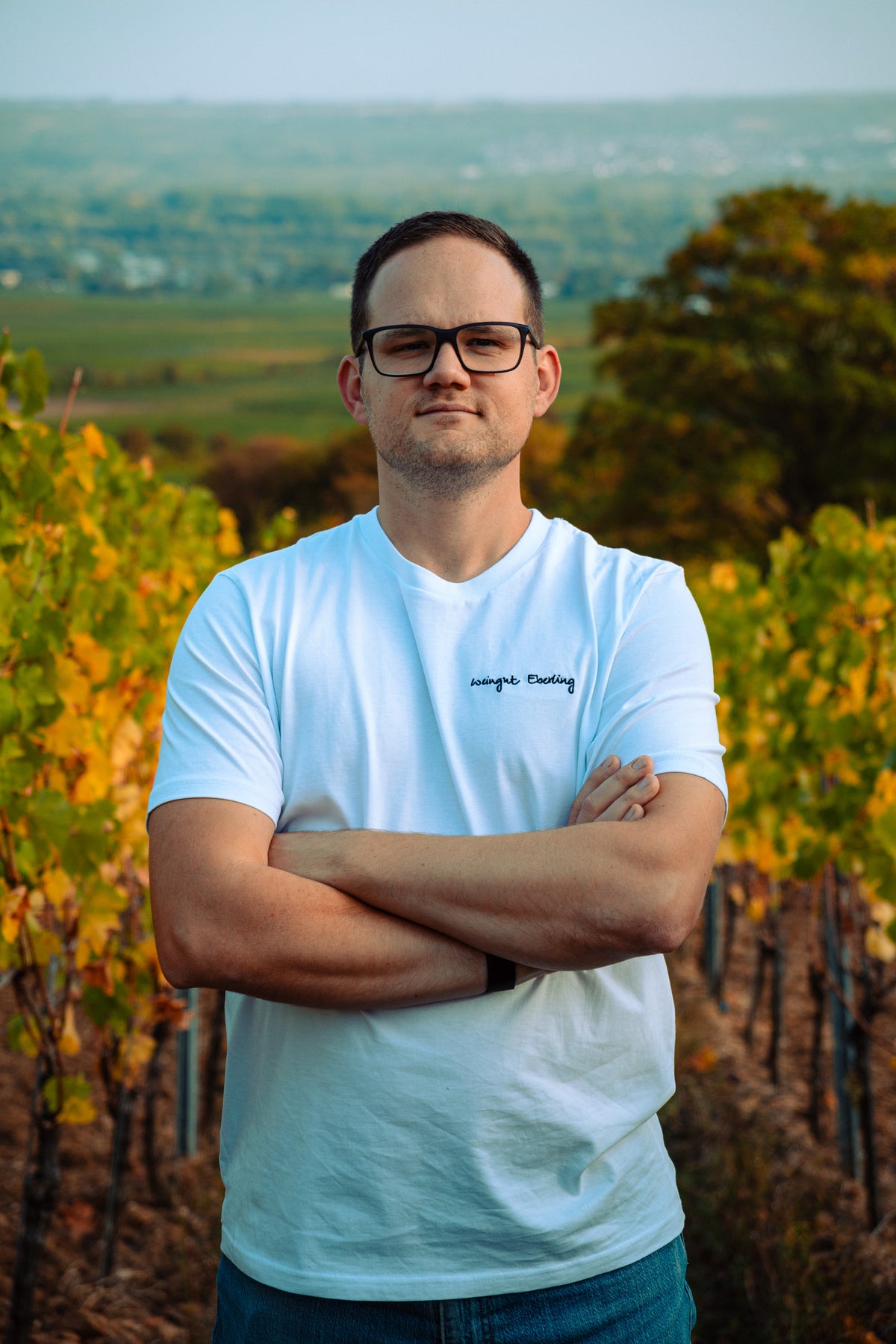 Mann im weißen „Weingut Eberling“-T-Shirt mit verschränkten Armen zwischen herbstlich gefärbten Rebzeilen, im Hintergrund der Blick ins Tal