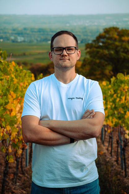 Mann im weißen „Weingut Eberling“-T-Shirt mit verschränkten Armen zwischen herbstlich gefärbten Rebzeilen, im Hintergrund der Blick ins Tal