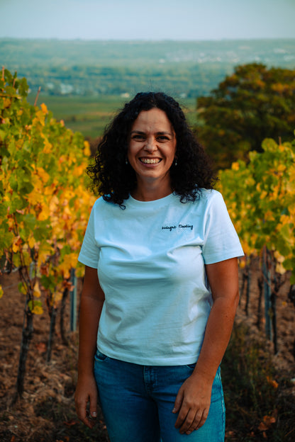Lächelnde Frau mit dunklen Locken im weißen T-Shirt mit gesticktem Schriftzug „Weingut Eberling“, im herbstlichen Weinberg